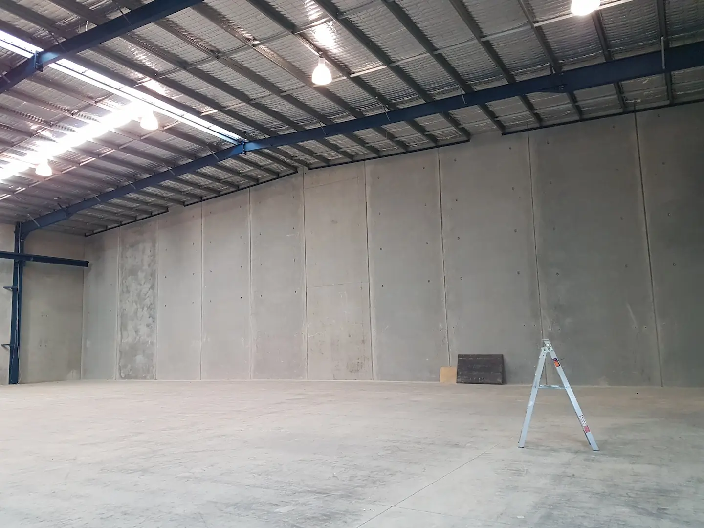 Interior of a spacious Brisbane storage facility showcasing the robust construction with concrete tilt-up panels. The industrial space is illuminated by natural light filtering through skylights and artificial light from high-bay fixtures. A metal step ladder is placed near the center, hinting at ongoing work or inspections.