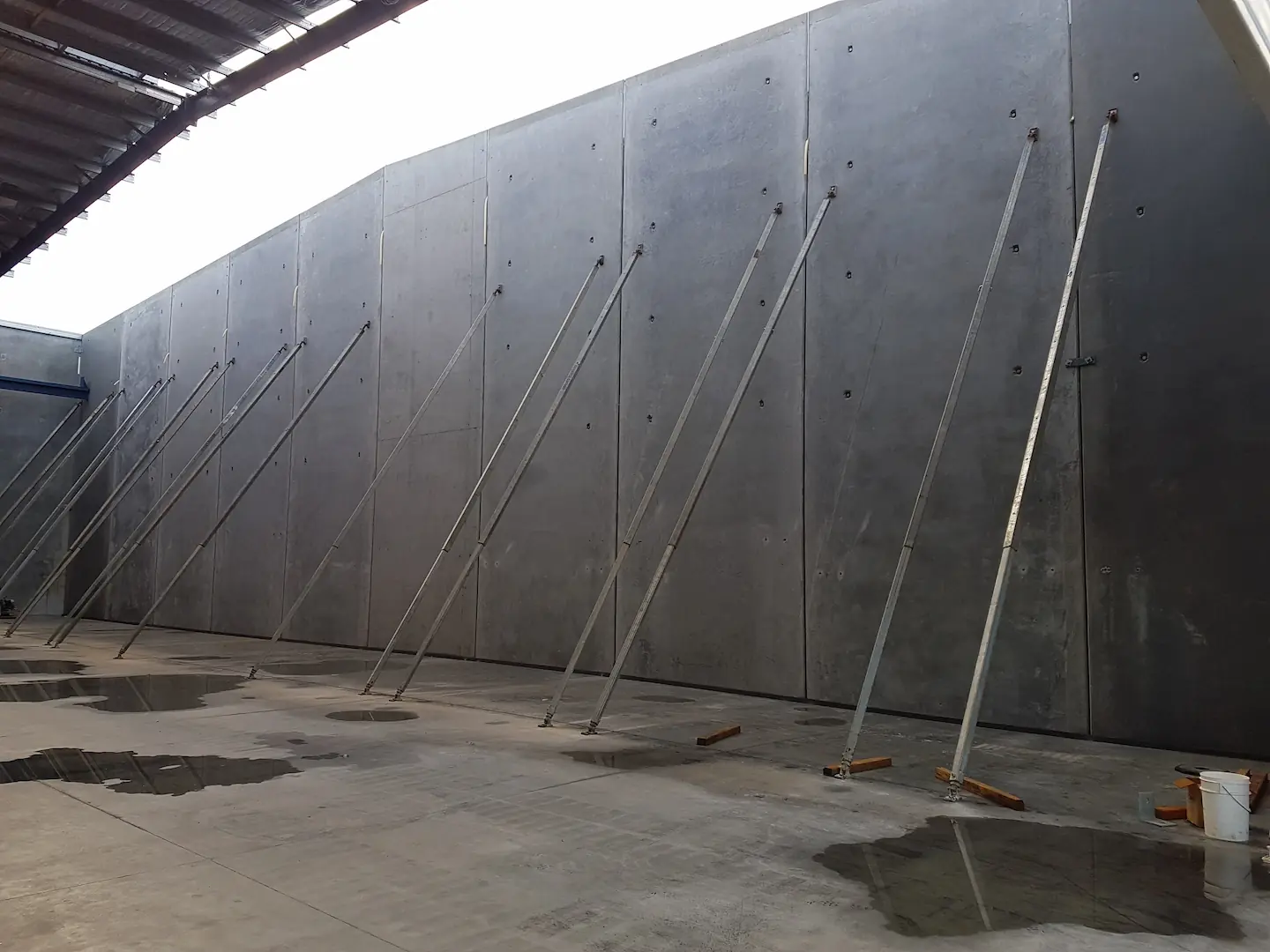 Concrete tilt-up panels being secured with braces in the construction of a Brisbane storage facility. The perspective highlights the scale and precision involved in erecting the massive panels. Reflections on the wet concrete floor suggest recent work or cleaning, and construction materials are scattered around, providing a glimpse into the construction process.