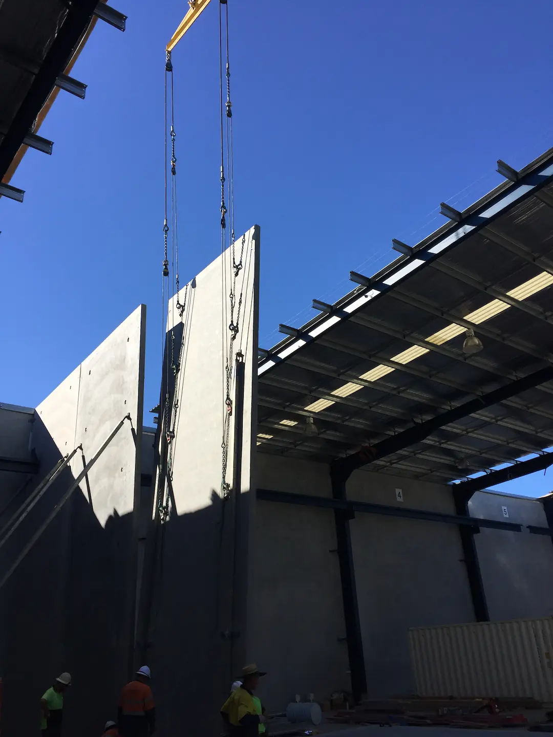 Construction workers oversee the precise lifting of a concrete tilt panel by a crane at the Brisbane storage facility site. The clear blue sky provides a contrasting backdrop to the industrial scene, emphasizing the panel's ascent. The dynamic of teamwork and engineering expertise is evident as the workers ensure the panel's safe and accurate placement.