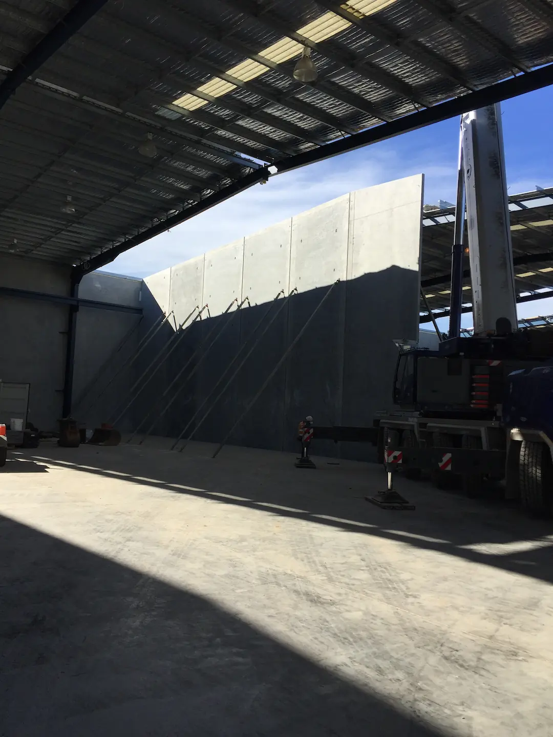 The interior of a commercial Brisbane storage facility under construction with a tall concrete tilt panel being supported by a mobile crane. The shadow of the structure is cast across the concrete floor, segmented by rays of sunlight entering from the open bay doors.