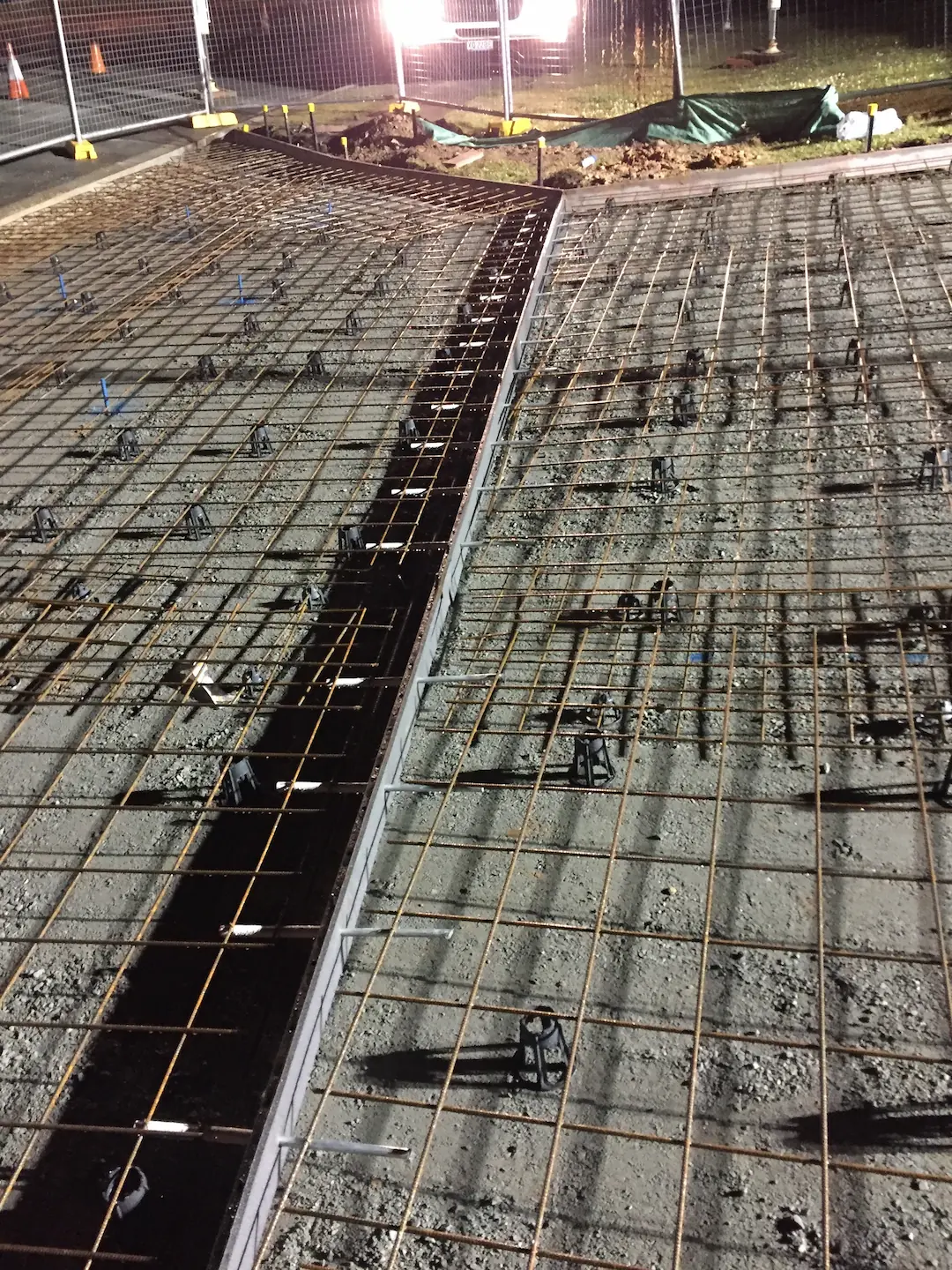 Preparation for a concrete foundation with metal reinforcement bars laid out and secured at night under artificial lighting.