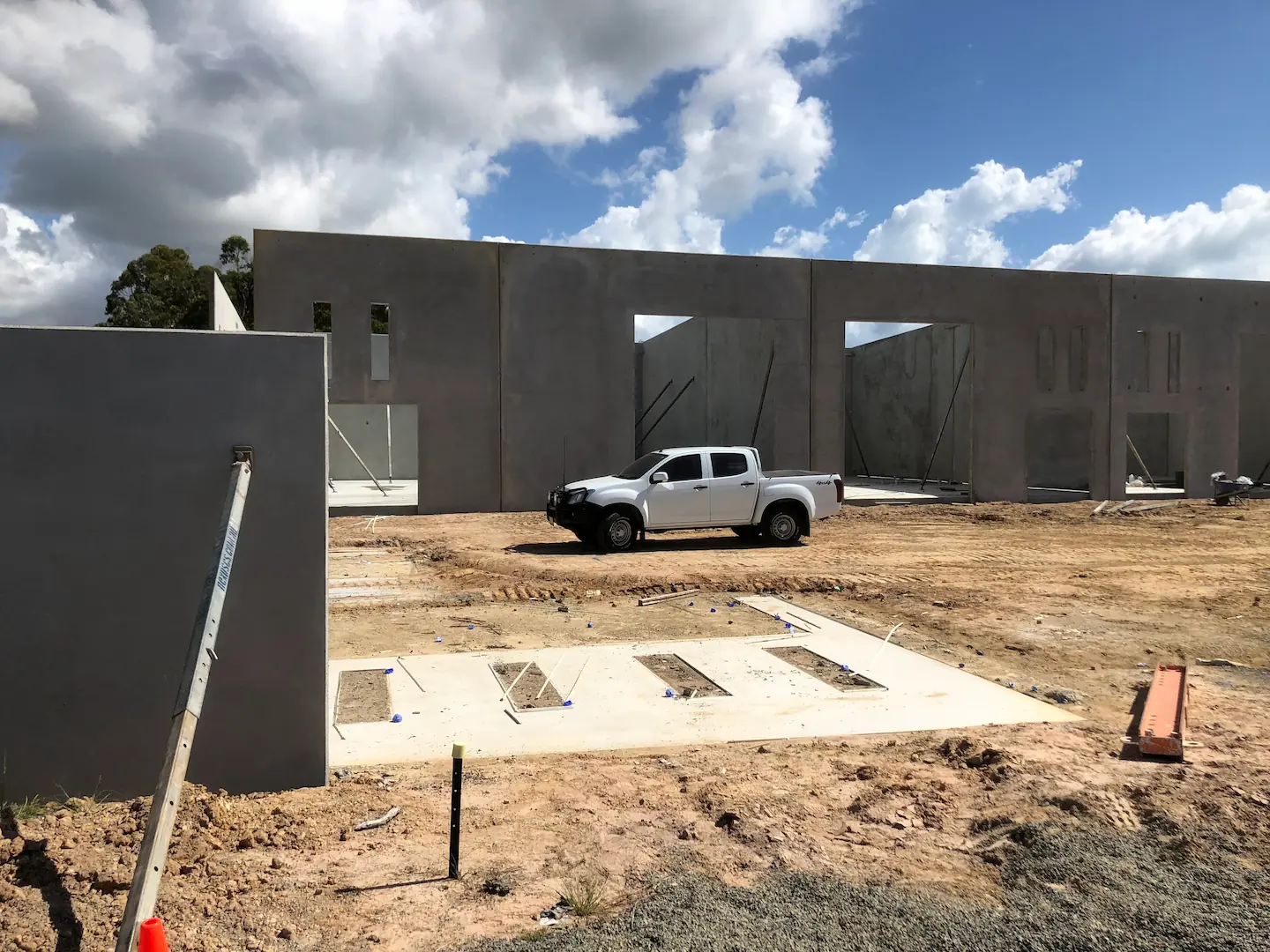  Early-stage construction of the Hervey Bay Industrial Complex showing several large concrete tilt panels propped up in an open field with bracing rods, under a cloudy sky. A white pickup truck is parked on site, providing scale to the massive panels.