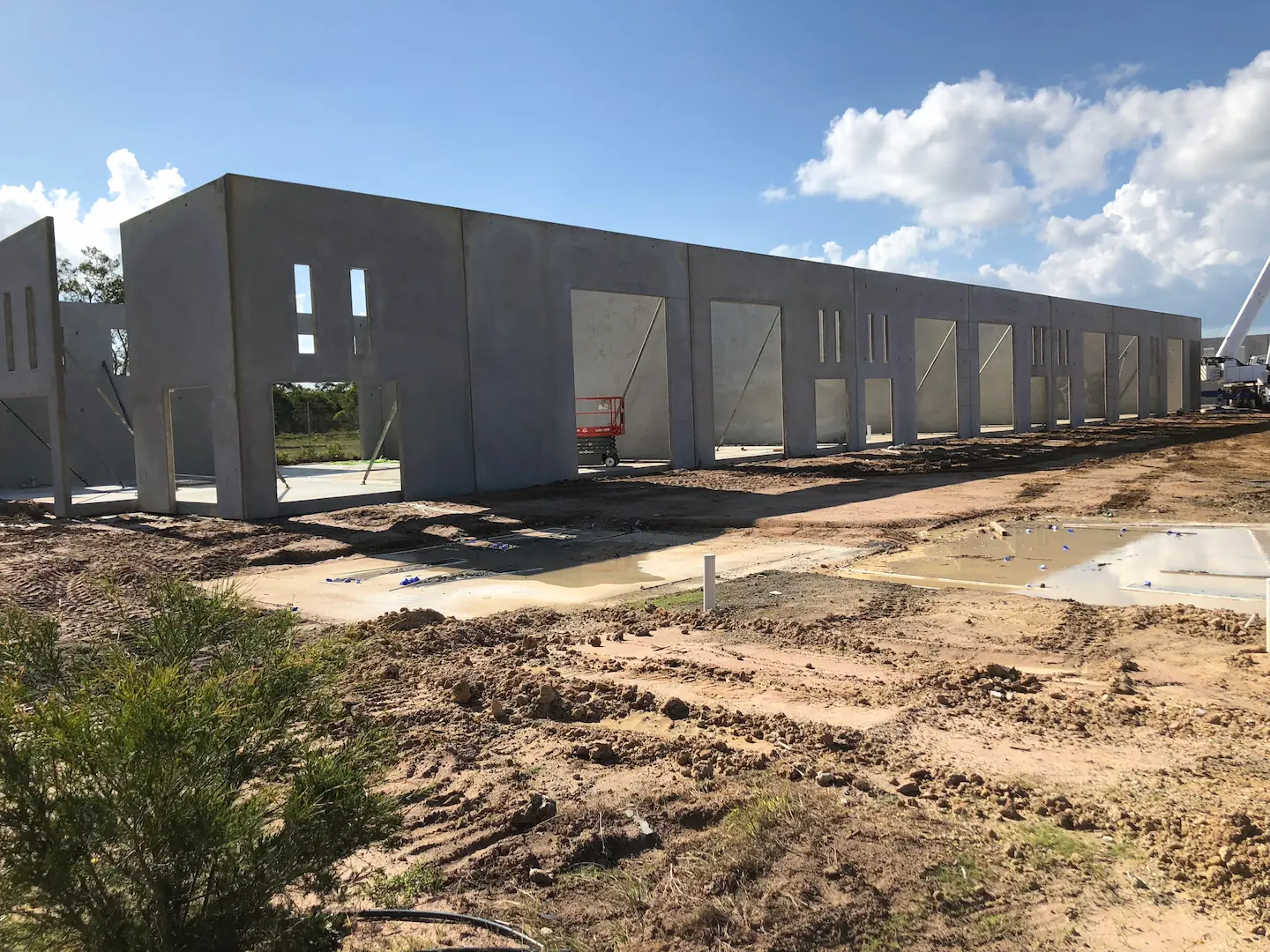 A series of tall concrete panels installed in a row at the Hervey Bay Industrial Complex construction site. The ground is muddy with puddles of water, reflecting the panels under a blue sky with fluffy clouds.