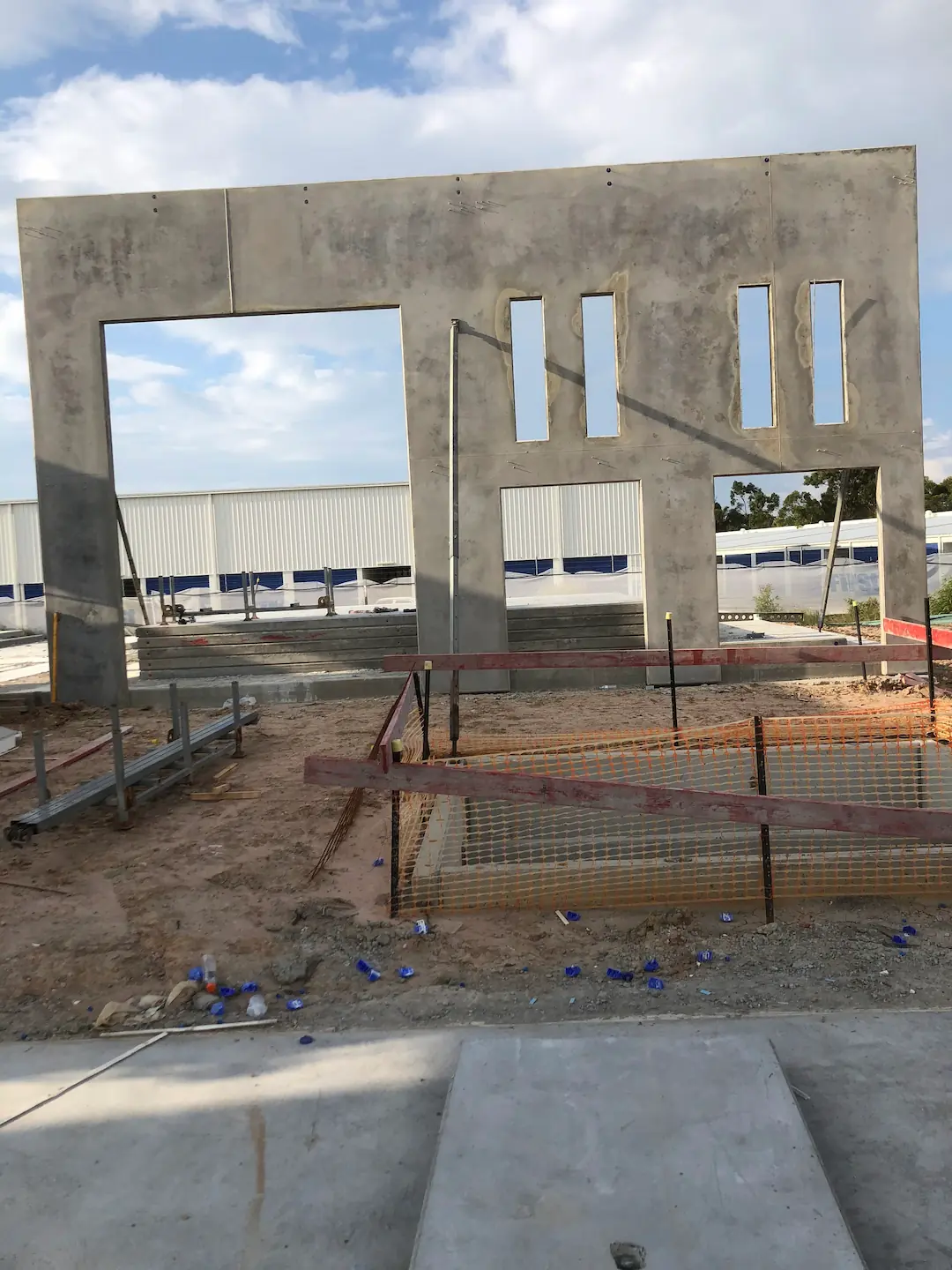 A standalone precast concrete panel with door and window openings stands at the Hervey Bay Industrial Complex site. The ground around is scattered with construction debris and rebar frames, under a partly cloudy sky. 