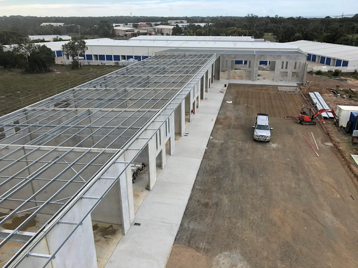 Elevated view of a construction site at Hervey Bay Industrial Complex with a steel roof framework in place above concrete walls. Construction machinery and a white truck are visible on the ground, indicating ongoing work.