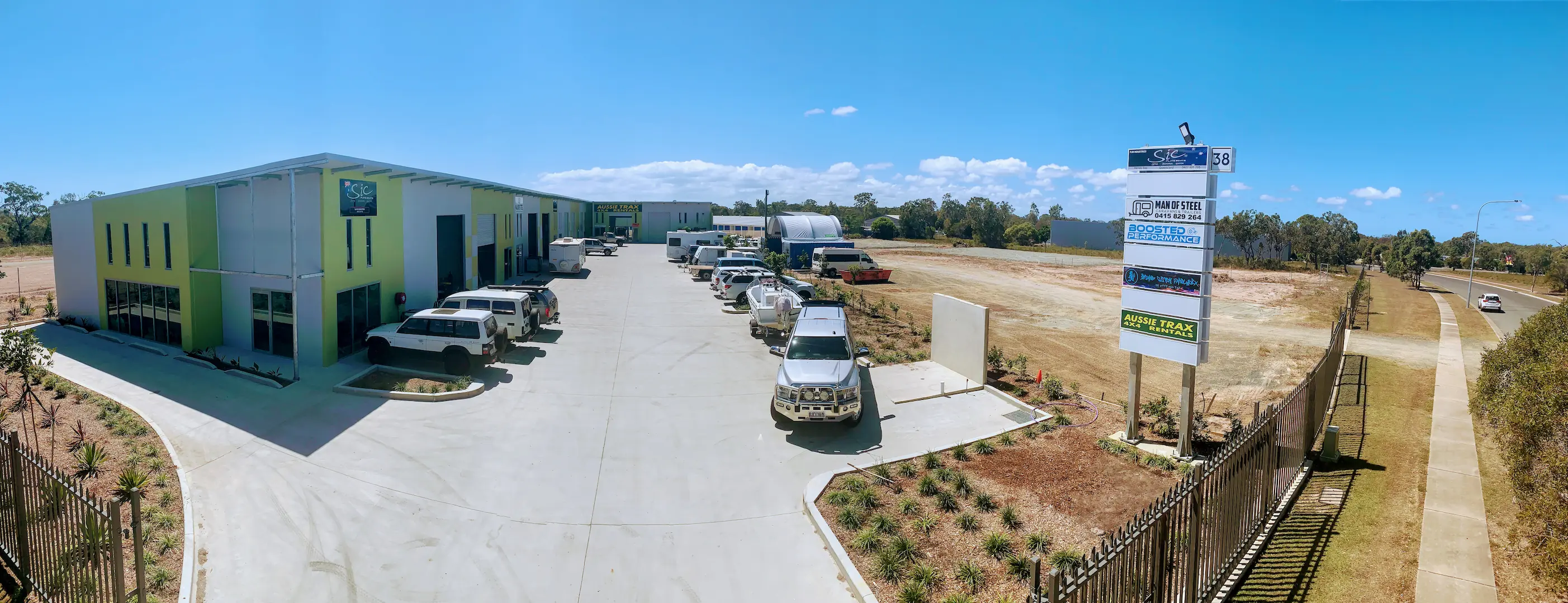 Panoramic view of the Hervey Bay Industrial Complex with a row of newly constructed light green commercial buildings. The complex features a paved central driveway with multiple vehicles parked, surrounded by landscaping and a vacant field extending towards a highway.