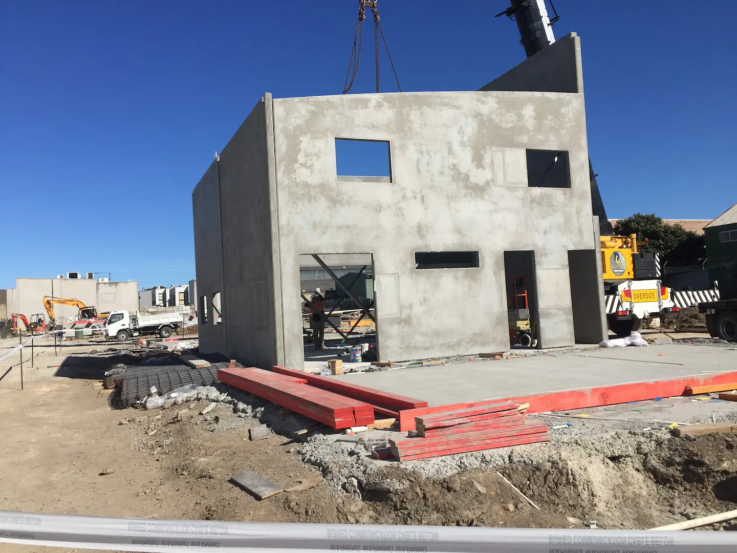 A concrete building with modern window cutouts stands with a nearly completed facade under a bright blue sky at the Kirawee project site, with construction materials and equipment foregrounding the scene.