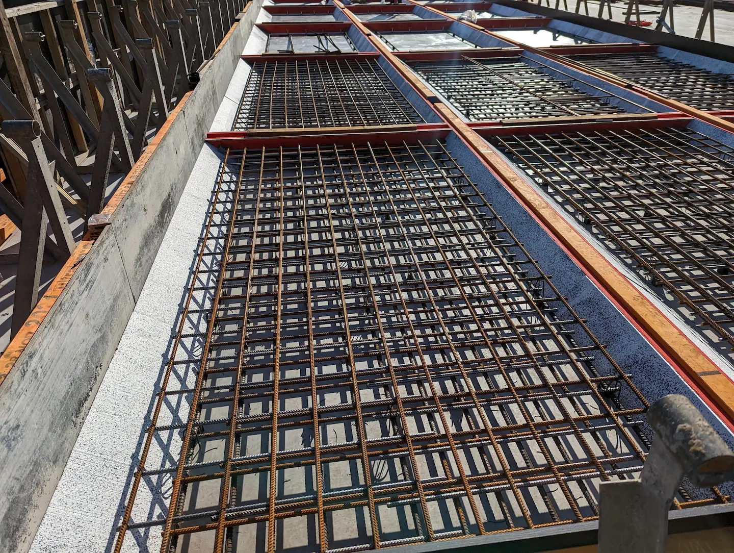 Overhead view of reinforced steel rebar grids in a formwork setup for a concrete slab at a construction site, with a white concrete edge visible and formwork structures in the background under clear skies.