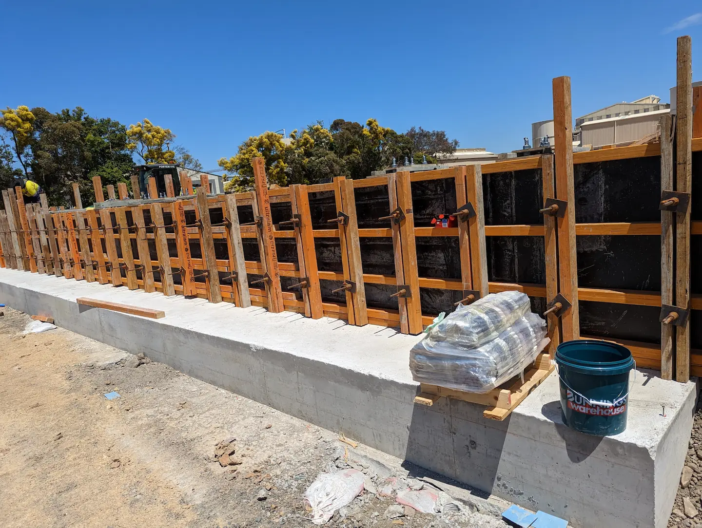 Daytime construction scene showing a concrete footing with vertical rebar and wooden formwork in preparation for a pour, with construction materials and a blue bucket on site, set against a clear sky with trees in the background.