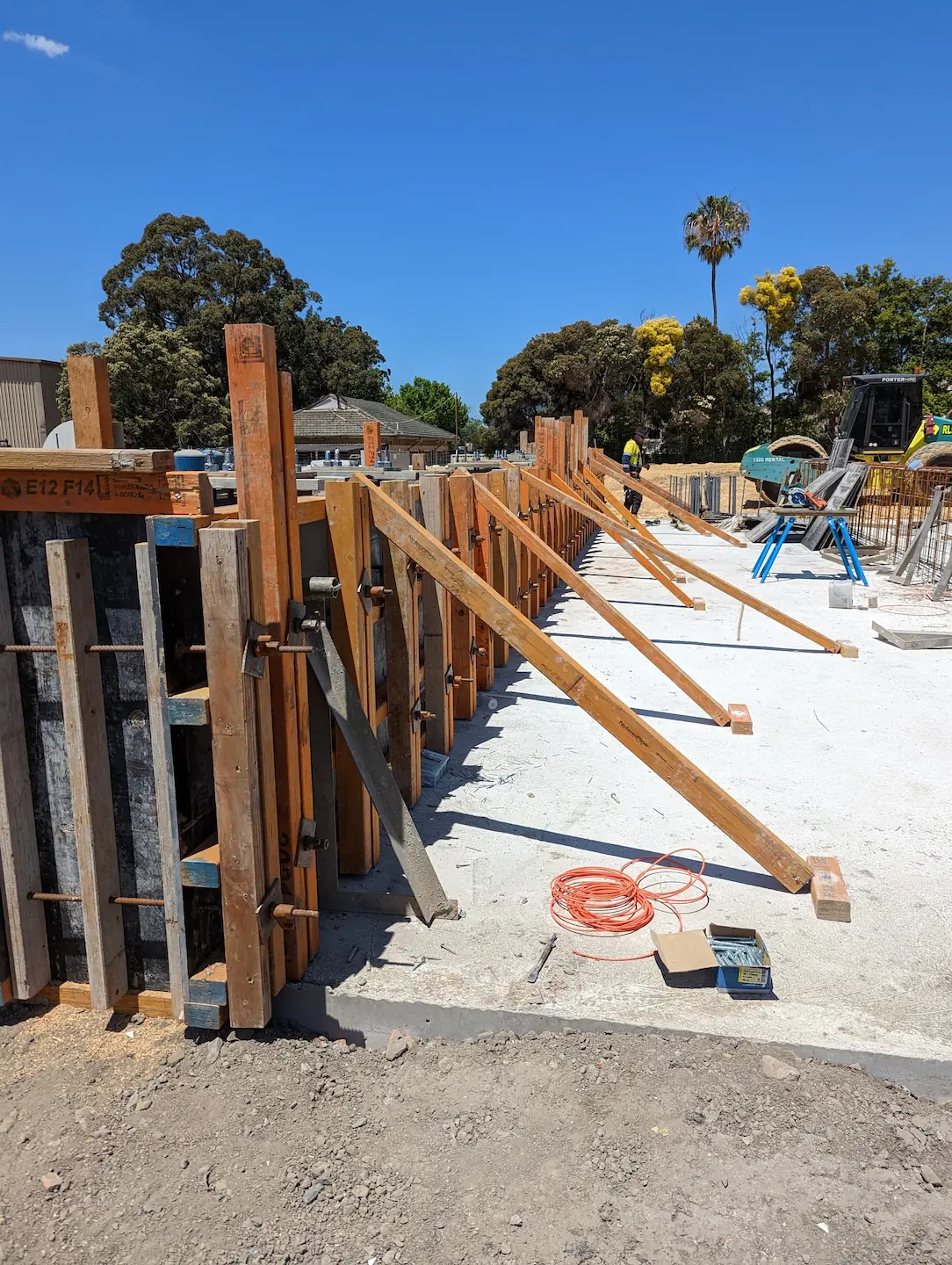 A construction site with complex wooden formwork supporting steel reinforcing bars in preparation for a concrete pour. An orange electrical cable, scale, and construction tools are visible on the freshly laid concrete, set against a sunny backdrop with flora and construction equipment.