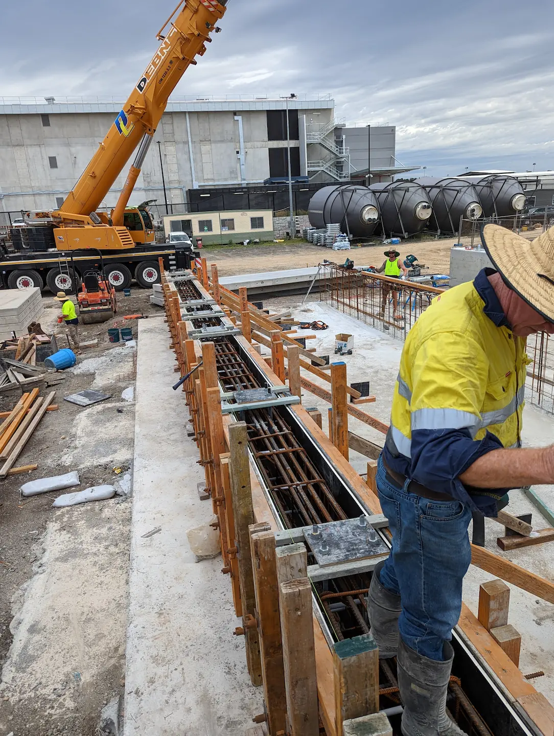 Construction workers engaged in meticulous prep work at a commercial concreting site, with deep trenches lined by wooden forms and steel reinforcement bars. A crane hovers in the background near storage tanks, highlighting the industrial environment and scale of the project.