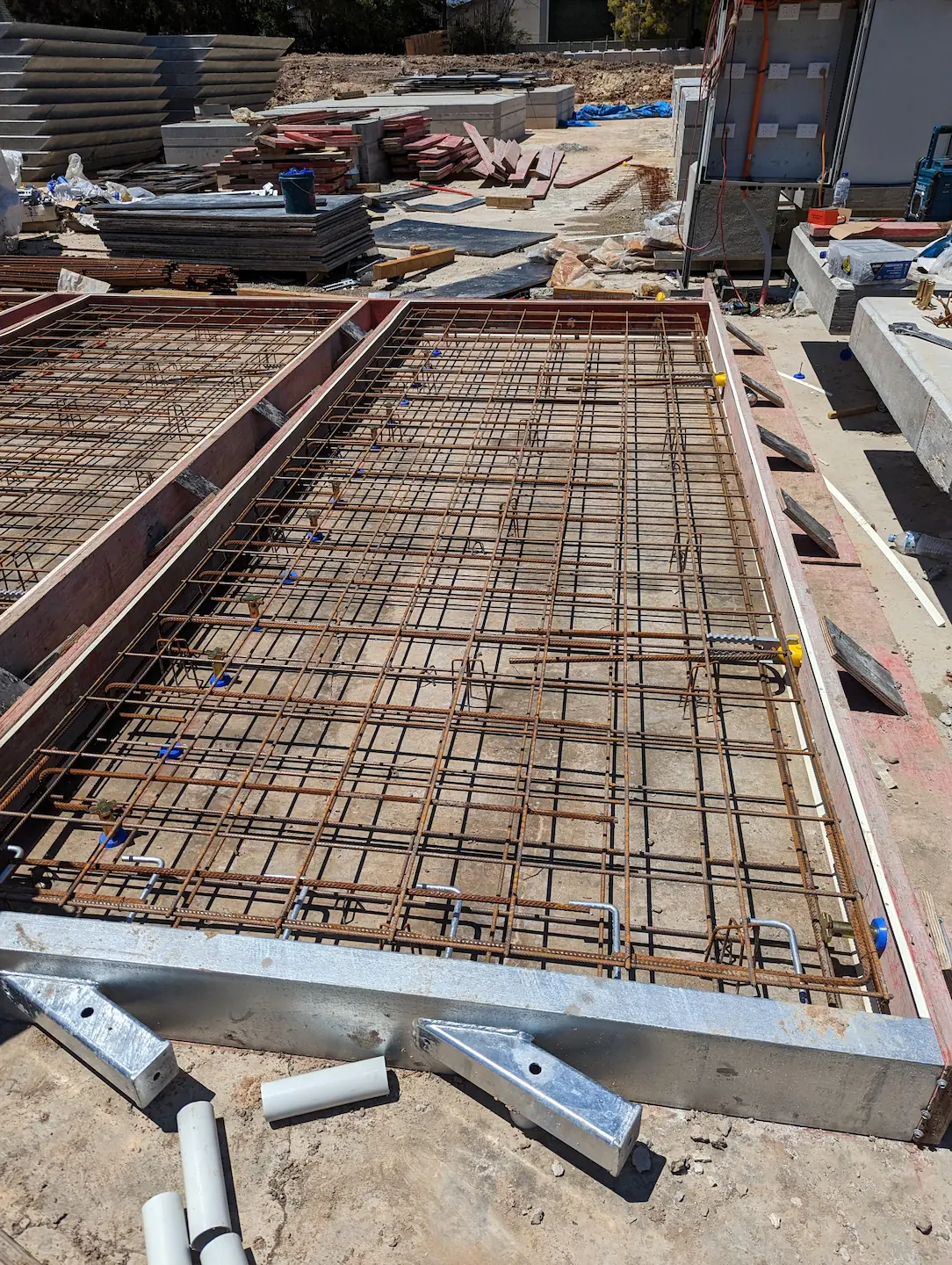 Detailed image of a concrete foundation formwork filled with an intricate grid of steel reinforcing bars, ready for concrete pour, with construction materials and site buildings in the background.