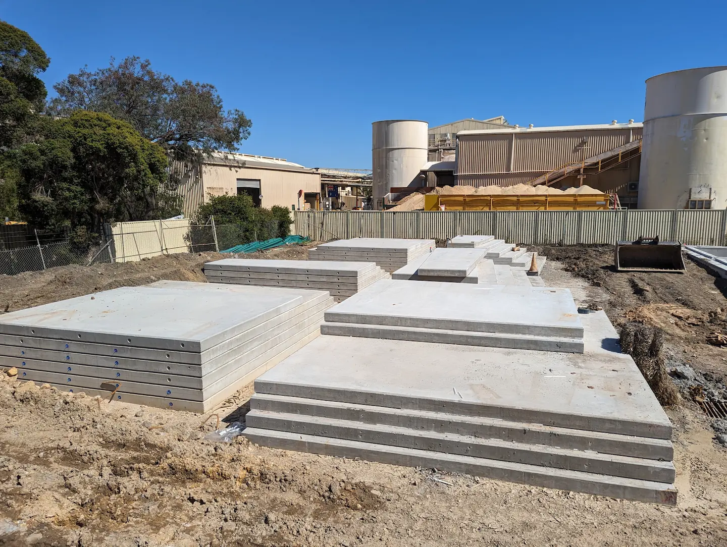 A series of precast concrete slabs are arranged in a staggered pattern on a construction site, with industrial tanks and buildings in the background, indicating preparation for installation in a large-scale commercial project.