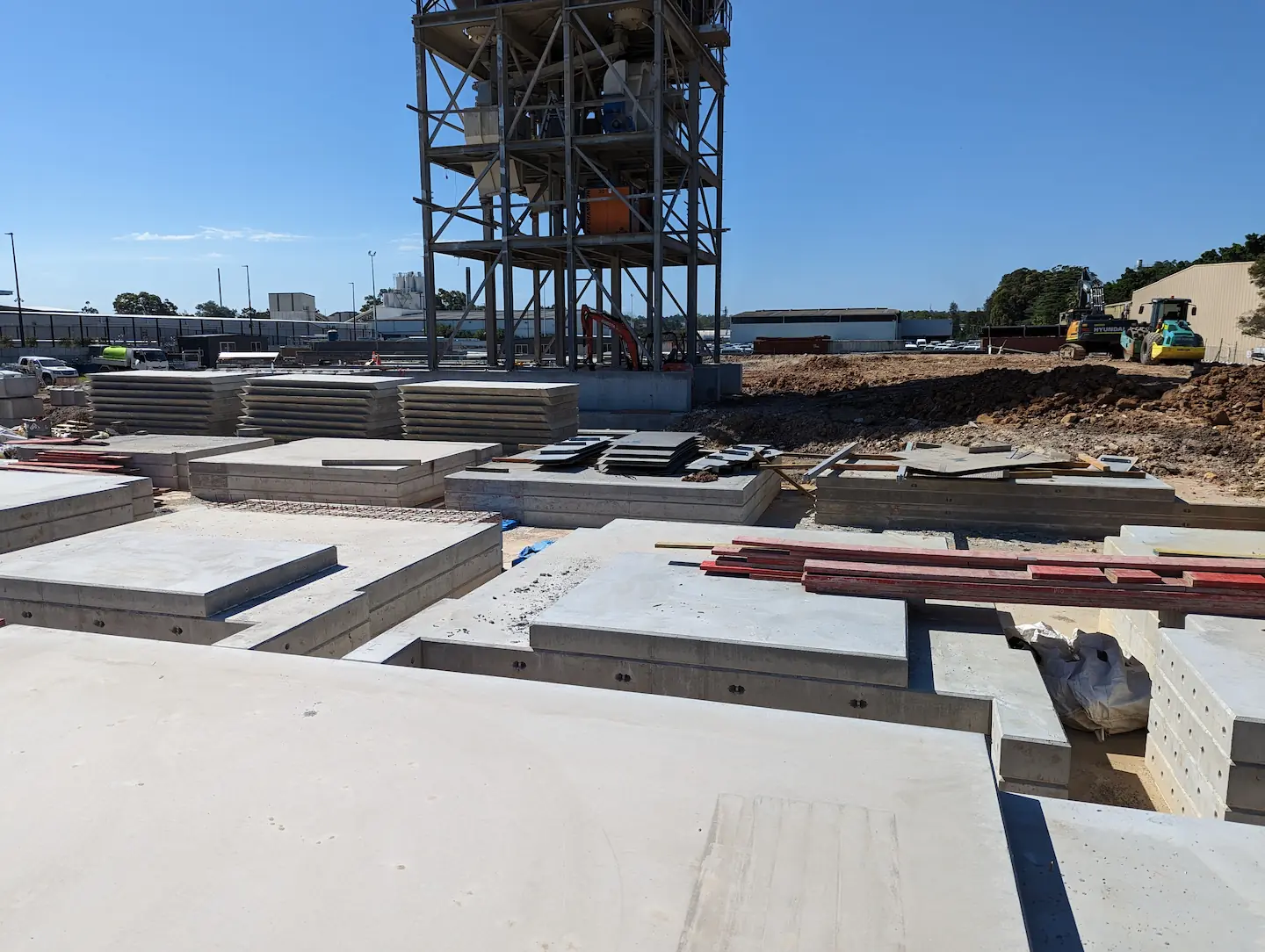 The photo shows a variety of precast concrete panels arranged horizontally on a construction site, with a backdrop of industrial buildings and vegetation. The panels are in different stages of installation, with visible steel reinforcing and anchoring elements, reflecting the meticulous preparation for constructing a robust structure.