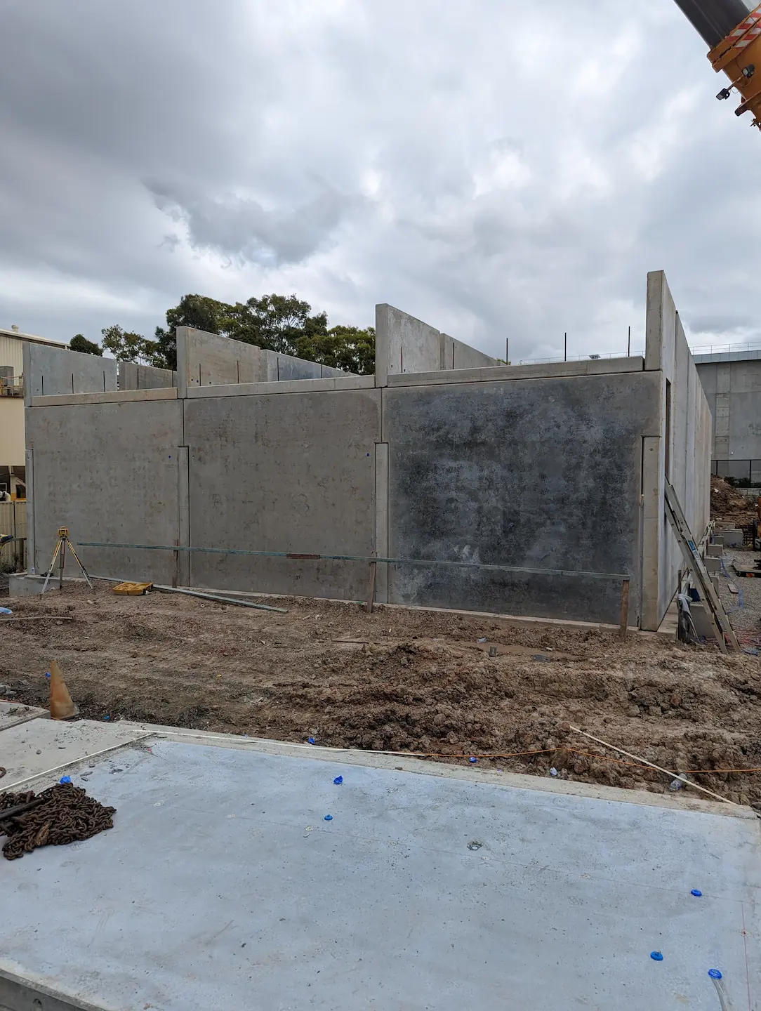 This image captures a cloudy sky above the concrete walls of a construction site. The foreground shows concrete foundations with protruding reinforcement bars and wooden stakes, hinting at the early stages of a significant construction project, with the promise of complex architecture to come.